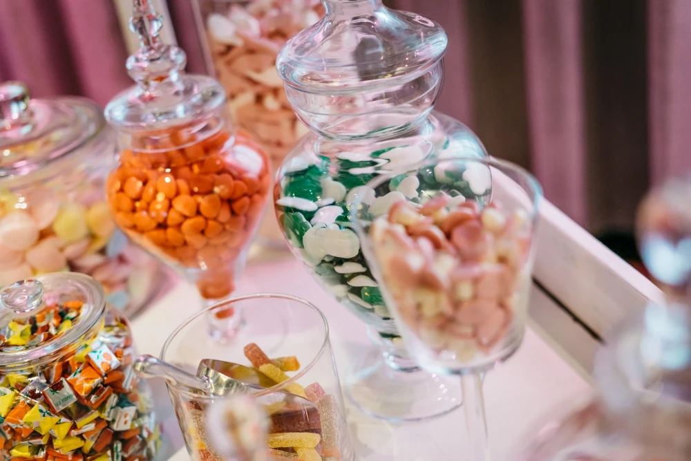 Verschiedene Glasgef&auml;&szlig;e gef&uuml;llt mit bunten S&uuml;&szlig;igkeiten und Bonbons auf einem wei&szlig;en Tablett, dekoriert mit rosa Vorh&auml;ngen im Hintergrund. Die Beh&auml;lter haben unterschiedliche Formen und Gr&ouml;&szlig;en und sind mit orange, gr&uuml;n-wei&szlig;en und anderen farbigen Leckereien gef&uuml;llt.