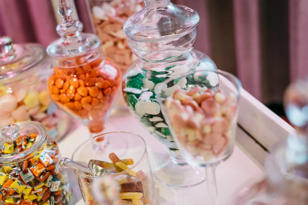 Verschiedene Glasgef&auml;&szlig;e gef&uuml;llt mit bunten S&uuml;&szlig;igkeiten und Bonbons auf einem wei&szlig;en Tablett, dekoriert mit rosa Vorh&auml;ngen im Hintergrund. Die Beh&auml;lter haben unterschiedliche Formen und Gr&ouml;&szlig;en und sind mit orange, gr&uuml;n-wei&szlig;en und anderen farbigen Leckereien gef&uuml;llt.