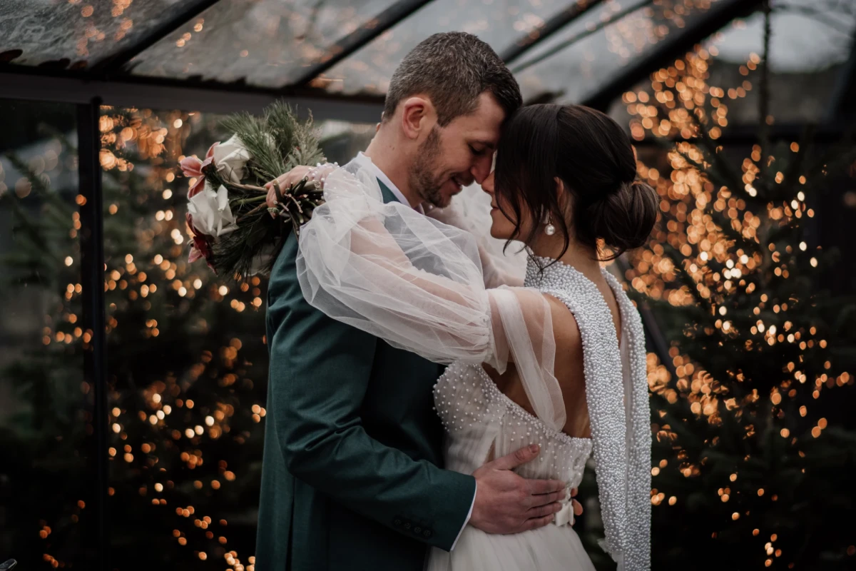 Brautpaar in z&auml;rtlicher Umarmung in einem Gew&auml;chshaus mit warmen Lichterketten im Hintergrund. Die Braut tr&auml;gt ein wei&szlig;es Spitzenkleid und einen Brautstrauss, der Br&auml;utigam einen dunkelgr&uuml;nen Anzug. Die Stimmung ist intim und romantisch.