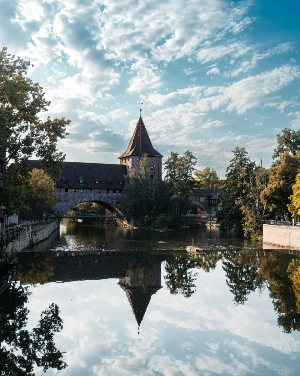 Historischer Stadtturm mit Br&uuml;cke spiegelt sich in ruhigem Wasser, umgeben von herbstlichen B&auml;umen und einem wolkigen Himmel. Die mittelalterliche Architektur der Steinmauer und des Turms wird durch die spiegelglatte Wasseroberfl&auml;che verdoppelt.