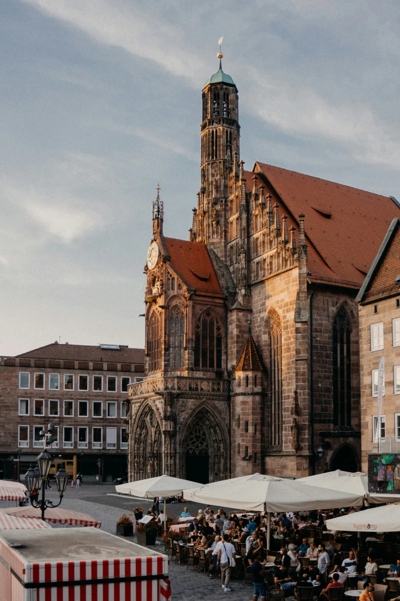 Kirche mit hohem Turm und rotem Dach auf einem belebten Platz in der Altstadt, umgeben von historischen Geb&auml;uden und Markst&auml;nden mit Menschen.