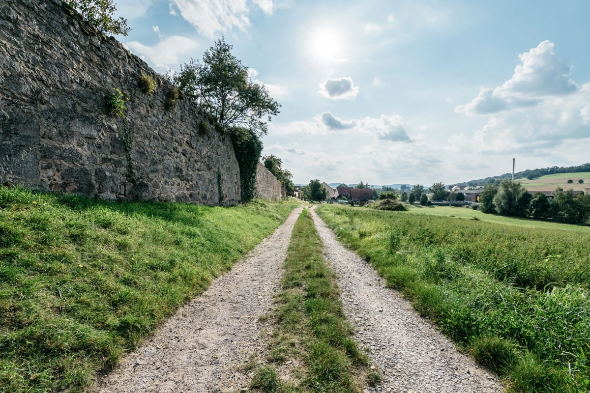Geschotterter Feldweg zwischen hoher Natursteinmauer und gr&uuml;ner Wiese, der sich durch eine l&auml;ndliche Landschaft mit Feldern und B&auml;umen im Hintergrund schl&auml;ngelt. Blauer Himmel mit leichten Wolken &uuml;berspannt die idyllische Szenerie.