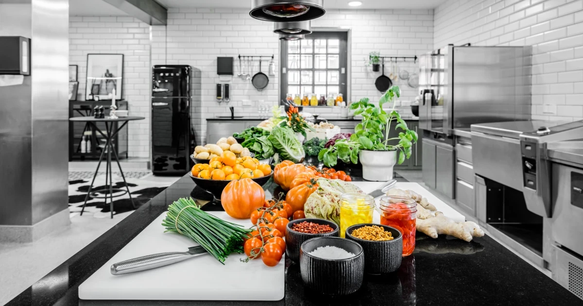 Frische K&uuml;rbisse, gr&uuml;ne Kr&auml;uter und verschiedene Gew&uuml;rze auf einem schwarzen K&uuml;chentisch in einer professionellen Edelstahl-Restaurantk&uuml;che mit wei&szlig;en Fliesenw&auml;nden und Industrieausr&uuml;stung.