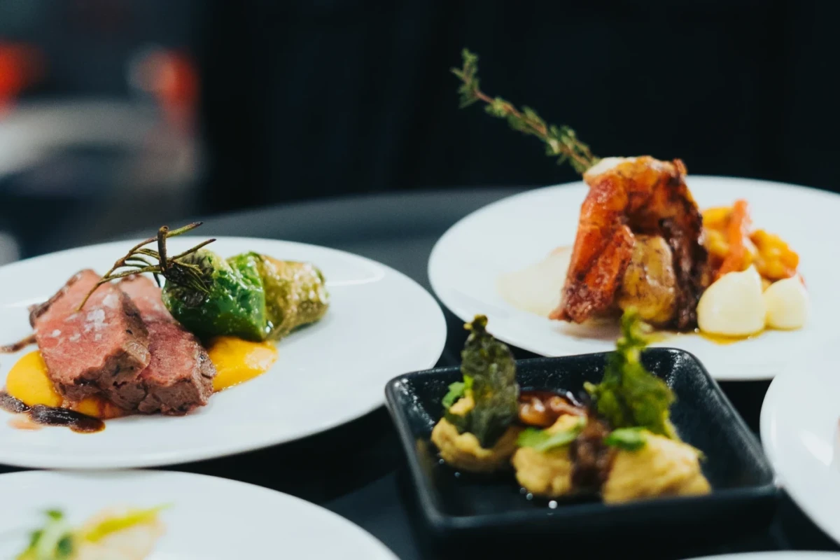 Drei elegant angerichtete Teller mit Gourmet-Hauptgerichten: Rosa gebratenes Fleisch mit gelber Sauce und gr&uuml;nem Gem&uuml;se, ein gebratenes Fleischgericht mit orangefarbener Sauce und ein weiteres Gericht auf einem schwarzen Teller, alle kunstvoll und professionell dekoriert.