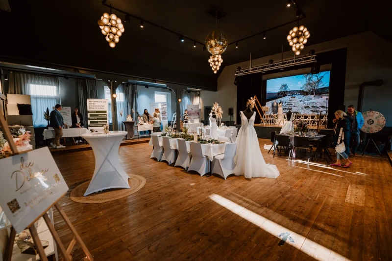 Hochzeitslocation mit eleganten, wei&szlig; bezogenen Tischen, h&auml;ngenden Kugelleuchten und einem Brautkleid, das auf einem St&auml;nder steht. Ein gro&szlig;er Bildschirm im Hintergrund zeigt eine Panoramaaufnahme, und der Raum hat einen warmen Holzboden mit ged&auml;mpftem Licht.