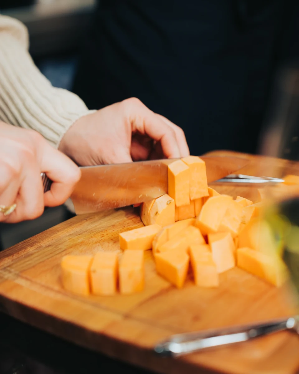 Auf einem Holzbrett werden K&uuml;rbis- oder Butternut-W&uuml;rfel von einer Hand mit einem Messer geschnitten, w&auml;hrend der Hintergrund verschwommen ist.