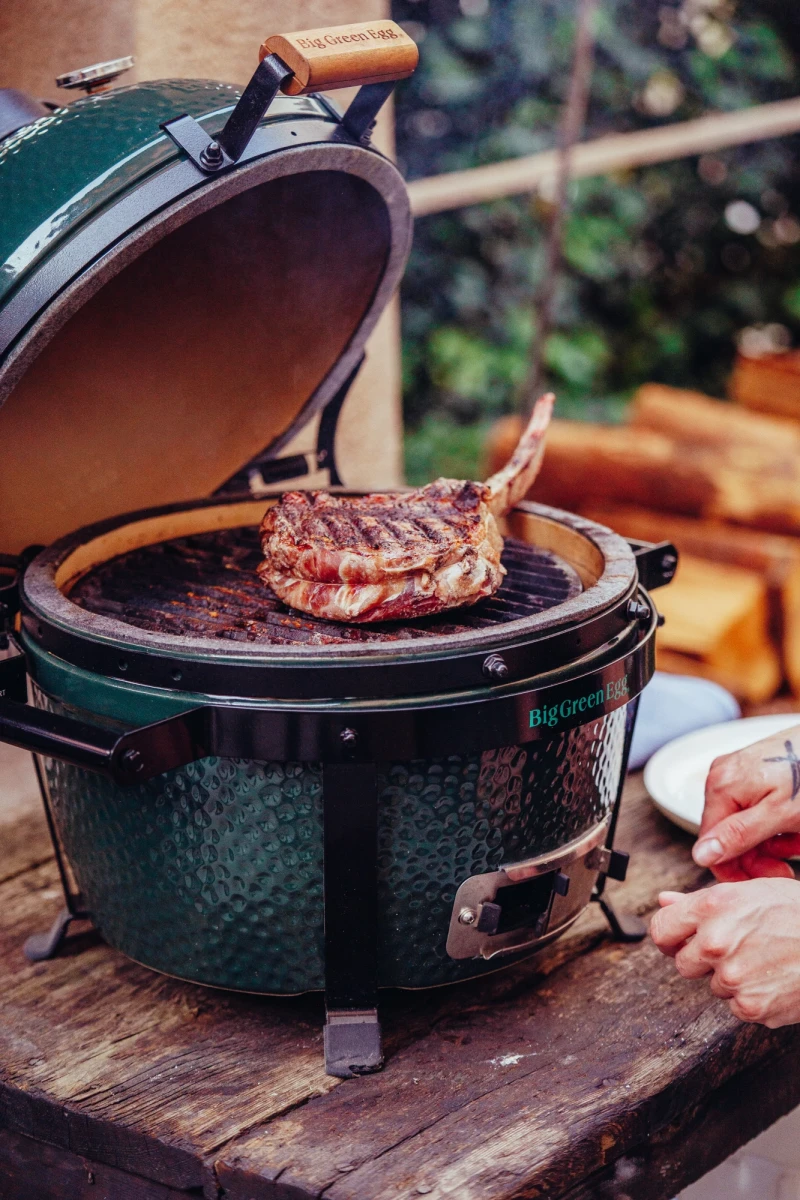 Saftig gegrilltes Tomahawk-Steak auf einem gr&uuml;nen Big Green Egg Keramikgrill, platziert auf einem rustikalen Holztisch mit verschwommenem Gartenhintergrund.