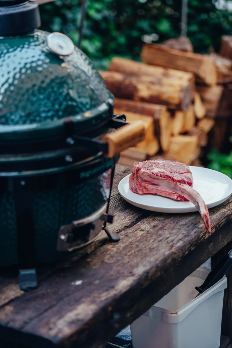 Rohes Rindfleisch auf einem wei&szlig;en Teller auf einem rustikalen Holztisch, neben einem gr&uuml;nen Keramikgrill und einem Stapel Holzscheite im Hintergrund.