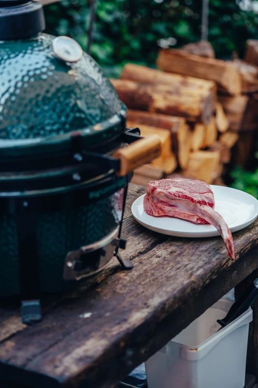 Rohes Rindfleisch auf einem wei&szlig;en Teller auf einem rustikalen Holztisch, neben einem gr&uuml;nen Keramikgrill und einem Stapel Holzscheite im Hintergrund.