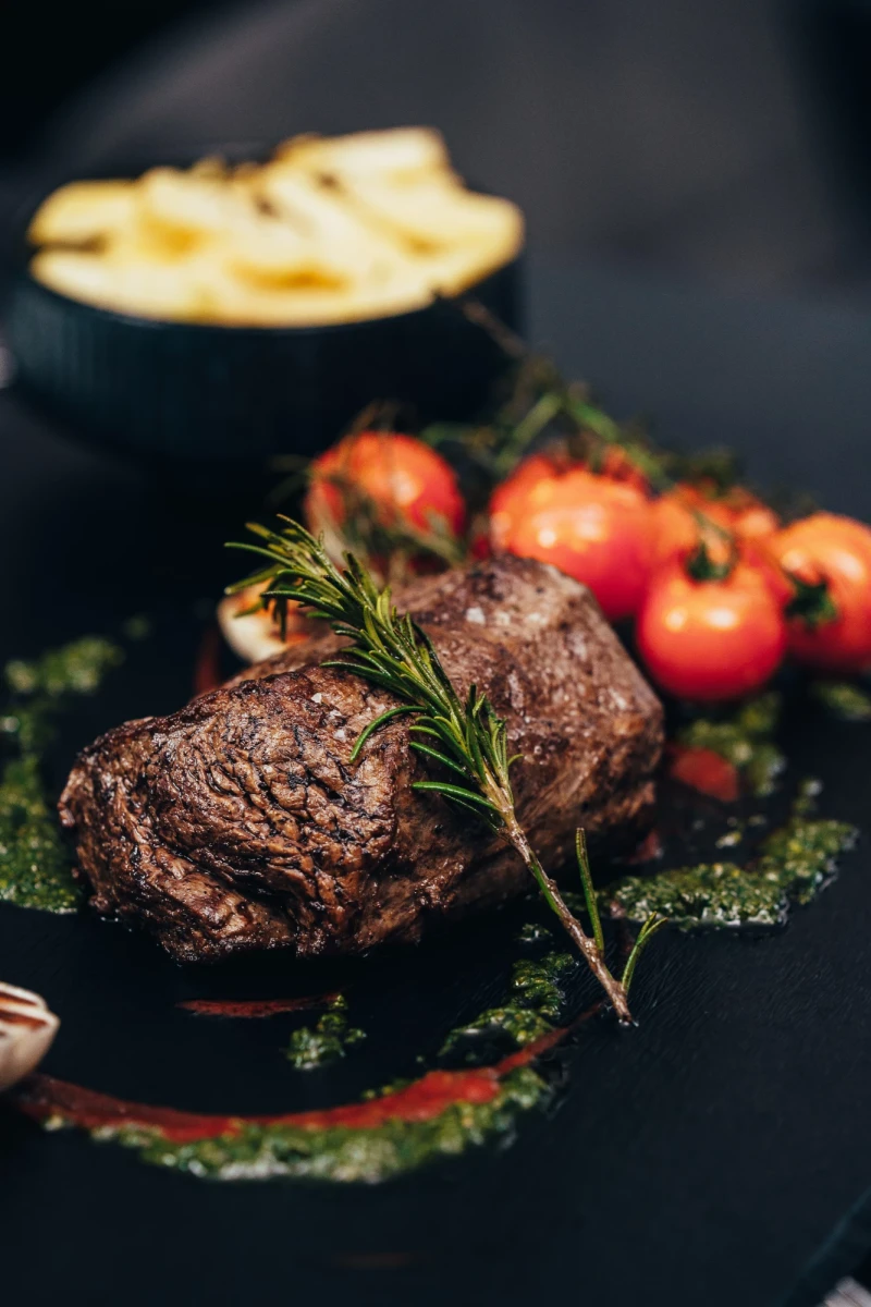 Zwei saftig gebratene Rinderfilets auf einem dunklen Teller mit Rosmarin, umgeben von roten Tomaten und einem gr&uuml;nen Kr&auml;uterrand. Im Hintergrund eine Schale mit goldbraunen Pommes frites.