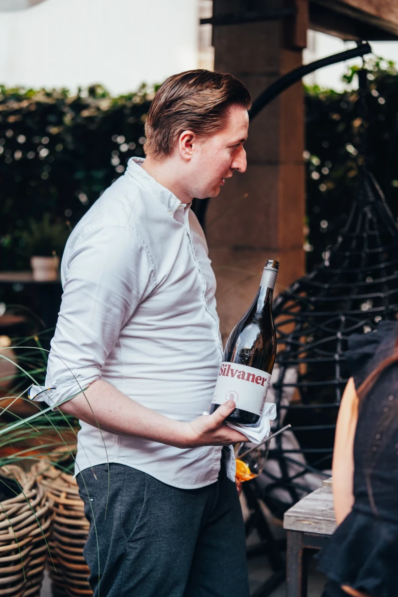 Mitarbeiter in wei&szlig;em Hemd steht auf einer Terrasse und h&auml;lt eine Weinflasche, w&auml;hrend er sich l&auml;chelnd nach rechts wendet, im Hintergrund sind verschwommene Pflanzen und Gartenm&ouml;bel zu sehen.