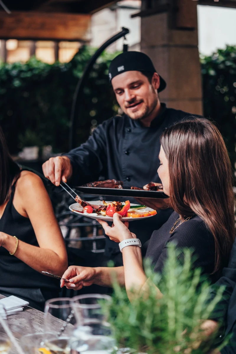 Koch in schwarzer Uniform und M&uuml;tze serviert eine Platte mit Speisen an einen Gast auf einer Terrasse mit Pflanze im Vordergrund.