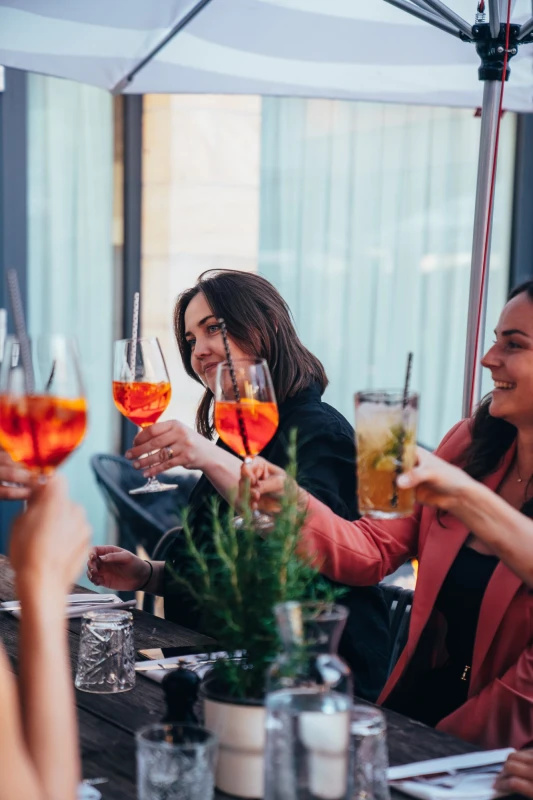 Zwei Frauen prosten sich mit orangefarbenen Cocktails in einem hellen Innenraum zu. Auf dem Tisch stehen Gl&auml;ser und Pflanzen, und sie scheinen in entspannter Stimmung zu sein.