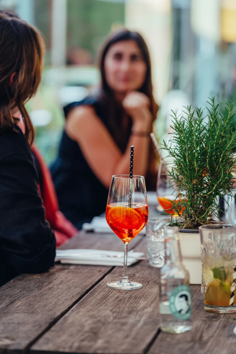 Auf einem Holztisch steht ein Aperol Spritz neben einem Rosmarin-Zweig und einem Getr&auml;nkeglas. Im Hintergrund sitzt eine Person in einem unscharfen Gastronomiebetrieb.