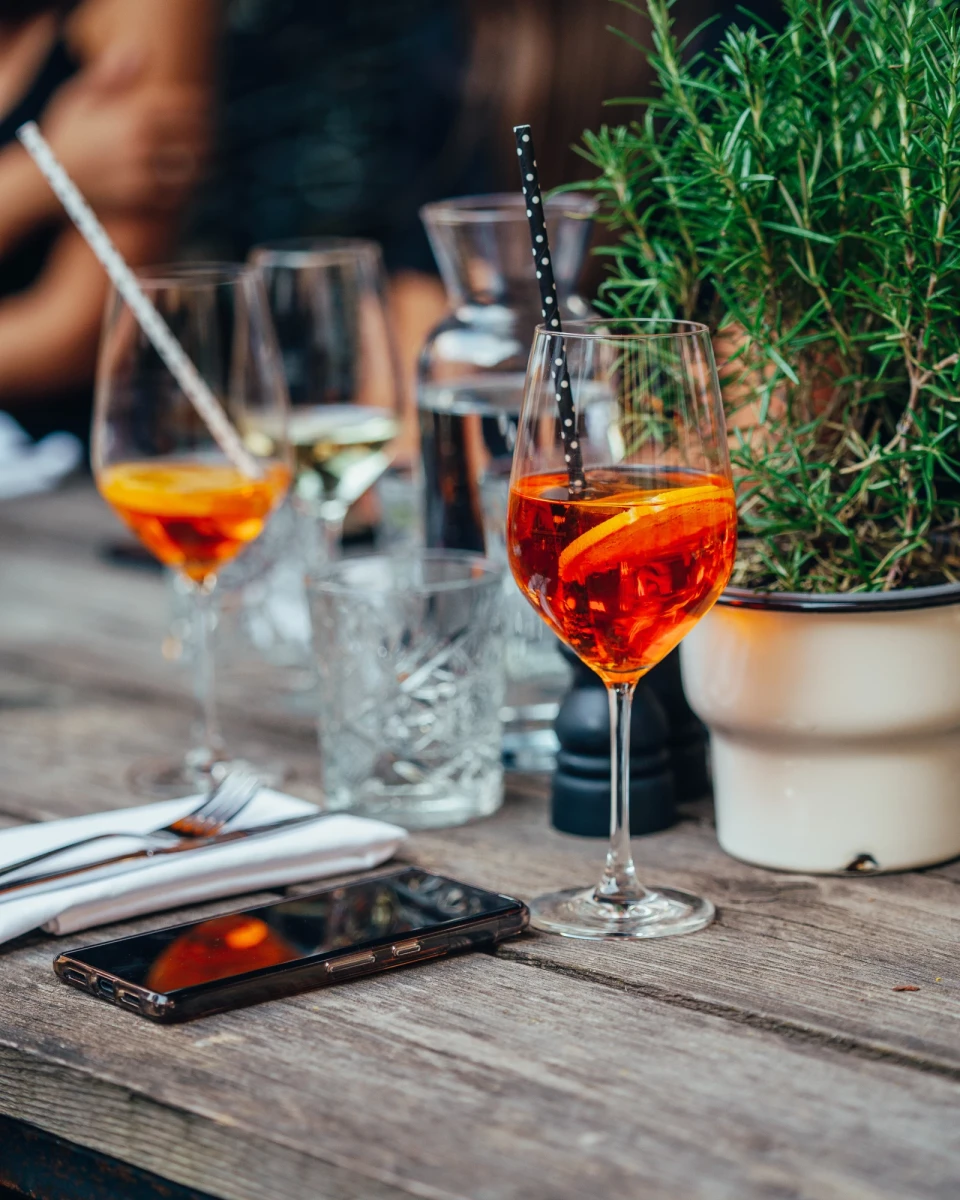 Zwei Cocktails in Weingl&auml;sern auf einem Holztisch, daneben eine gr&uuml;ne Pflanze und ein Smartphone. Die orangefarbenen Getr&auml;nke haben einen Barkeeper-Stil mit Metallst&auml;bchen und stehen in einer gem&uuml;tlichen Au&szlig;enumgebung.