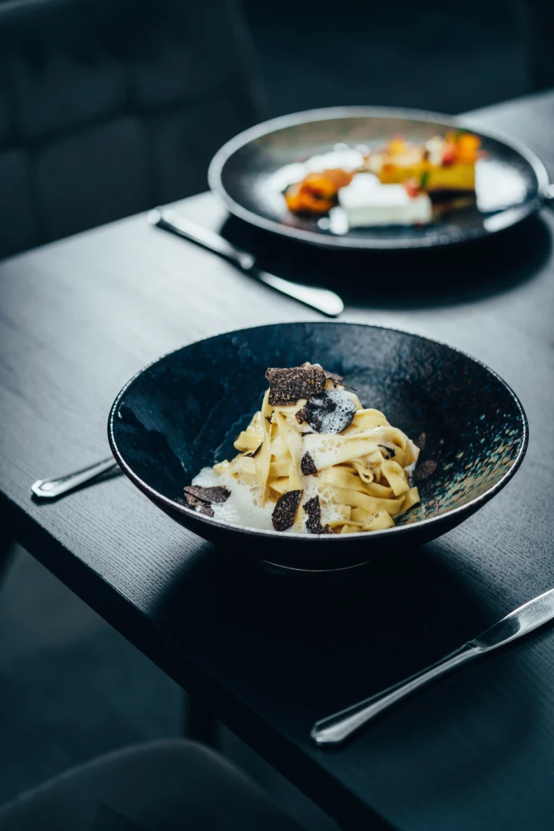 Cremefarbene Bandnudeln mit schwarzen Tr&uuml;ffelscheiben in einer dunklen Schale, serviert auf einem gedeckten Tisch mit silbernem Besteck und einer verschwommenen zweiten Schale im Hintergrund.