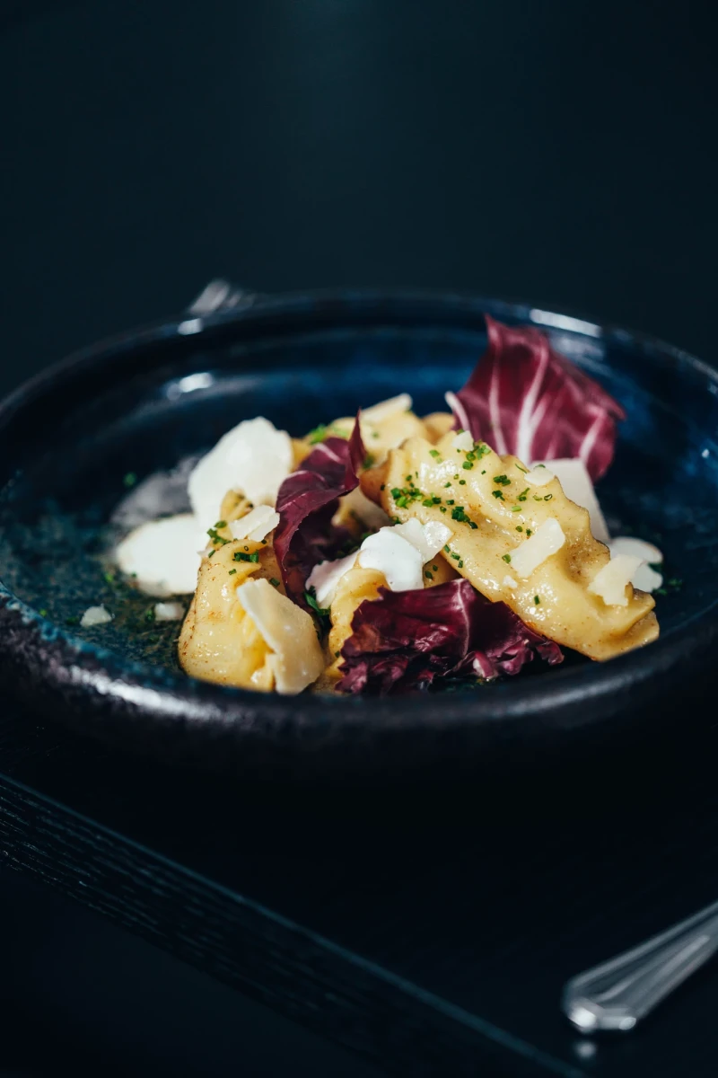 Tortellini mit wei&szlig;er Sauce auf dunkelblauem Teller, garniert mit roten Radicchio-Bl&auml;ttern und bestreut mit gehackten Kr&auml;utern, in einem eleganten, dunklen Ambiente serviert.