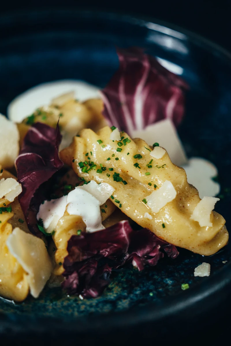 Kartoffelgnocchi mit cremigem K&auml;sesauce, bestreut mit gehackter Petersilie, serviert auf dunklem Teller mit violetten Radicchio-Bl&auml;ttern und br&ouml;ckeligen K&auml;sest&uuml;cken.