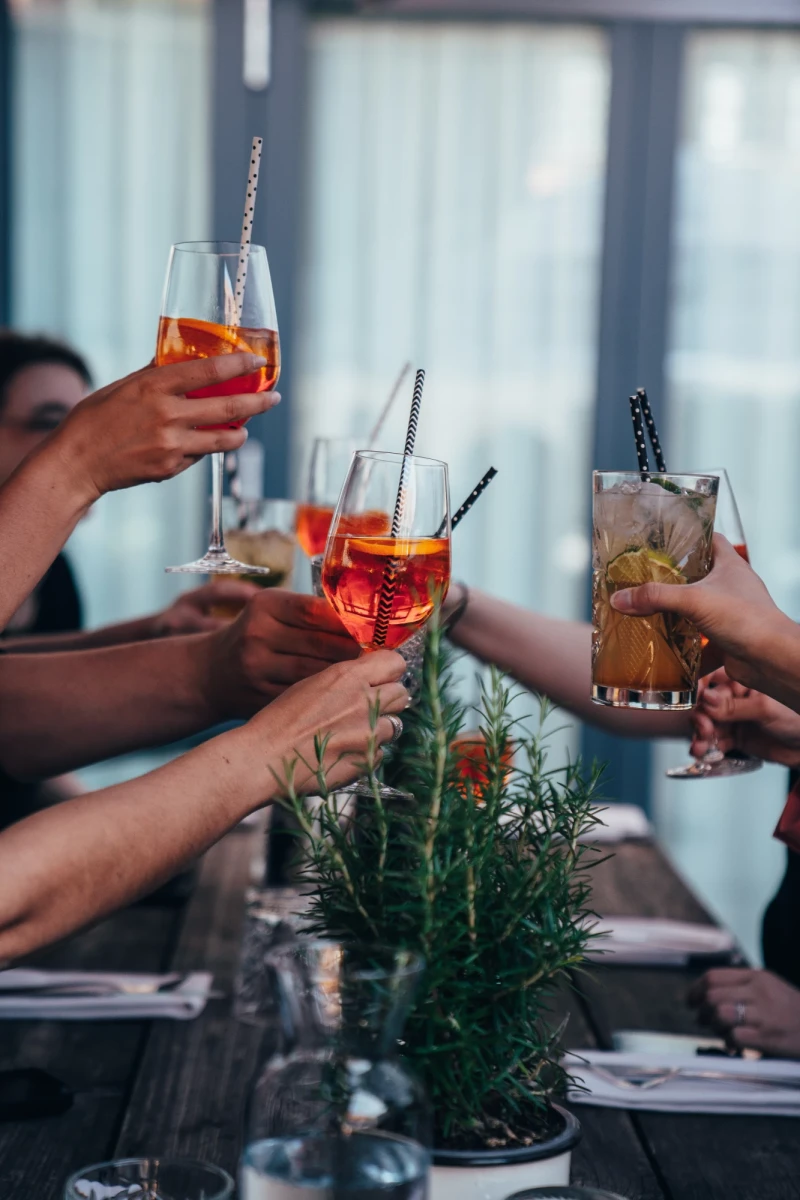 Mehrere H&auml;nde sto&szlig;en mit bunten Cocktails an einem Tisch mit Rosmarin an. Die Getr&auml;nke sind orangefarben und rot, serviert in eleganten Gl&auml;sern mit Metalltrinkhalmen vor einem unscharfen Hintergrund.