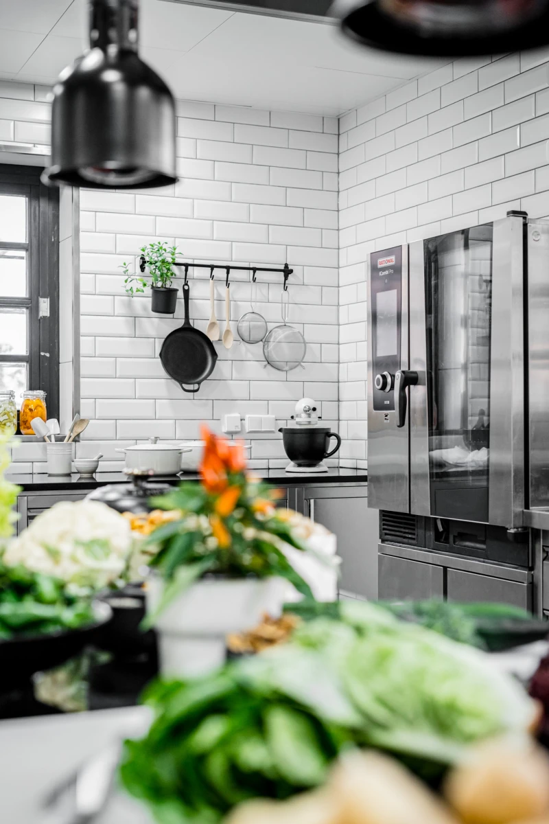 Moderne K&uuml;che mit wei&szlig;en Subway-Fliesen, Edelstahl-K&uuml;hlschrank und h&auml;ngenden T&ouml;pfen und Pfannen. Im Vordergrund liegen frische Gem&uuml;sesorten wie Kohl und Blumenkohl, mit einer Topfpflanze und Blumen im Hintergrund. Eine schwarze Deckenlampe und eine schwarze Tasse vervollst&auml;ndigen die minimalistische K&uuml;chenszene.