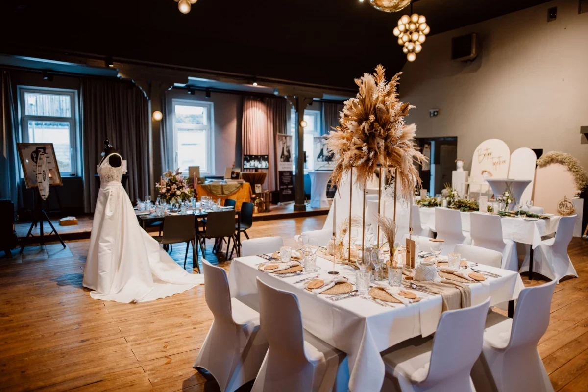 Festlich gedeckter Hochzeitstisch mit wei&szlig;er Tischdecke, goldenem Dekor und gro&szlig;er Pampasgras-Blumenarrangement in der Mitte, neben einem Brautkleid in einem eleganten Veranstaltungsraum mit Holzboden und Fenstern.