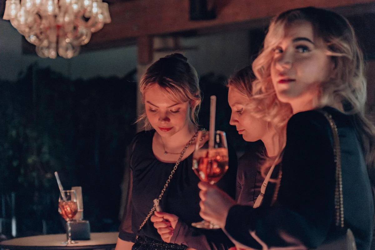 Drei junge Frauen in schwarzer Kleidung an einer Bar, eine Kronleuchter im Hintergrund. Sie trinken Cocktails und unterhalten sich in ged&auml;mpftem Licht, das eine warme, intime Atmosph&auml;re schafft.