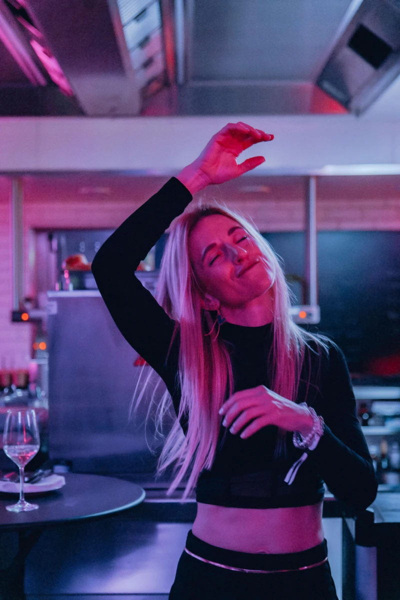 Eine Frau mit langen blonden Haaren tanzt l&auml;ssig in einer K&uuml;che mit rosa und blauen Neonlichtern, eine Hand &uuml;ber dem Kopf, ein Weinglas auf dem Tresen vor ihr.
