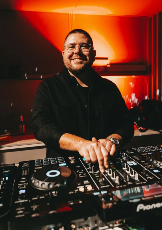 DJ an einem Mischpult in einem roten, atmosph&auml;rischen Raum. Er tr&auml;gt ein schwarzes Hemd und eine Brille und l&auml;chelt breit, w&auml;hrend er die Regler und Plattenteller bedient. Die Beleuchtung schafft eine warme, intensive Stimmung hinter ihm.