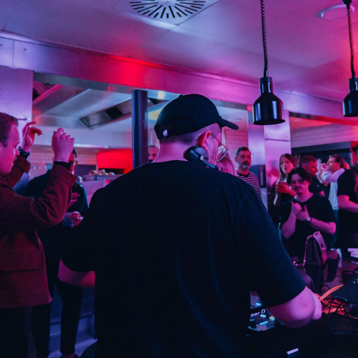 R&uuml;ckansicht eines DJs mit M&uuml;tze an einem Mischpult in einer belebten Bar mit rot-blauer Beleuchtung. Partyg&auml;ste tanzen und feiern im Hintergrund unter H&auml;ngelampen.