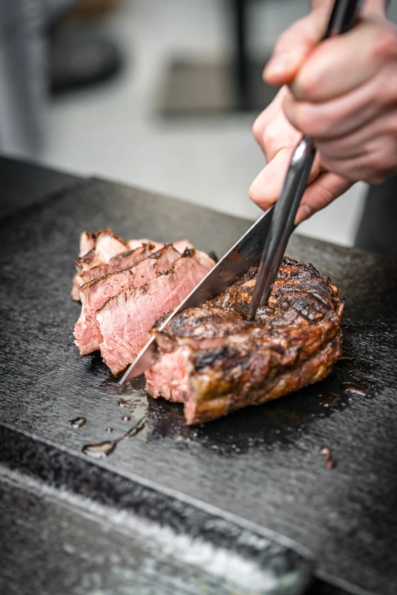 Gegrilltes Rindfleisch auf einer Steinplatte, eine Hand schneidet das zartrosa Steak mit einer Zange in Scheiben. Das Fleisch ist saftig und medium gebraten, mit einer gebr&auml;unten Kruste auf der Oberfl&auml;che.
