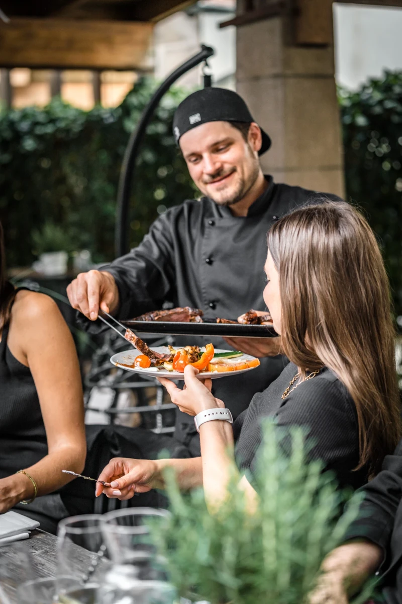 Ein Chefkoch in schwarzer Kochkleidung serviert einer Kundin eine Schale mit appetitlich angerichteten Speisen auf einer Terrasse mit Pflanzen im Hintergrund.