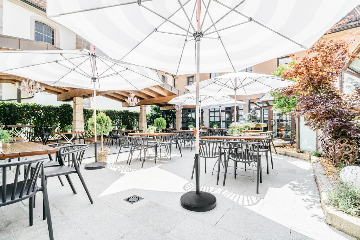 Heller, luftig gestalteter Au&szlig;enbereich eines Biergartens mit wei&szlig;en Sonnenschirmen, grauen Metalltischen und St&uuml;hlen. Bepflanzte Bereiche und Geb&auml;udeteile s&auml;umen den gepflasterten Sitzbereich, der eine moderne und einladende Atmosph&auml;re ausstrahlt.
