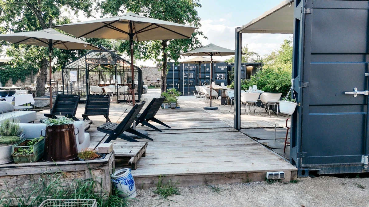 Moderne Terrasse mit grauen Liegen unter Sonnenschirmen, holzgepflasterte Fl&auml;che mit Sitzgelegenheiten und einem grauen Container im Seitenbereich, umgeben von B&auml;umen und Pflanzen.