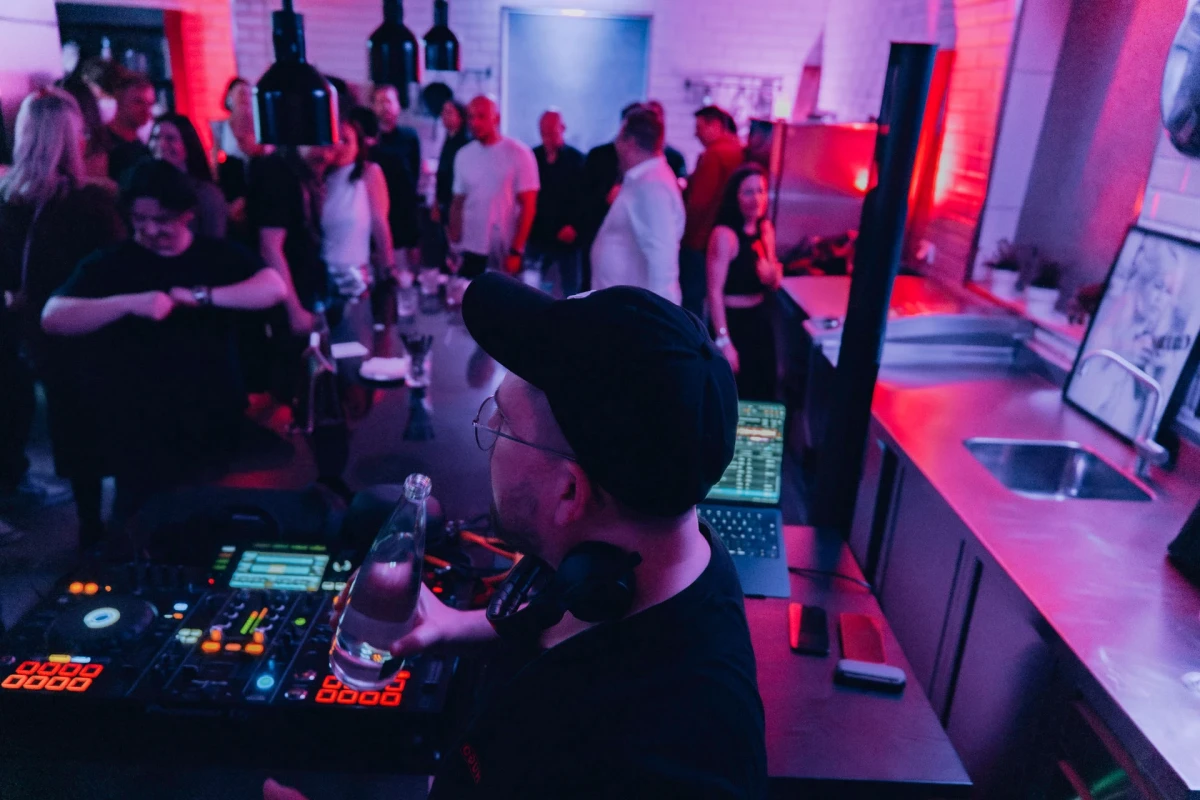 DJ in lebhafter Eventk&uuml;che mit G&auml;sten und bunten Lichtern.