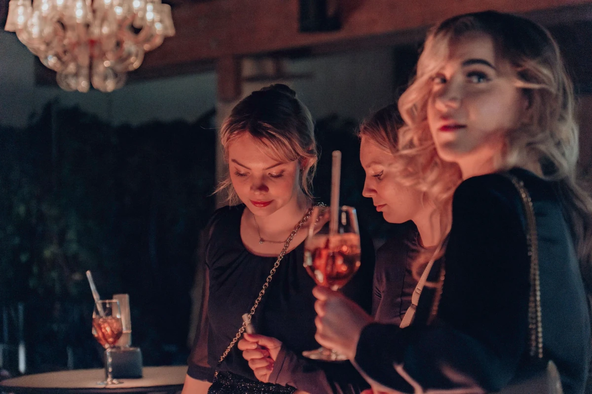 Drei junge Frauen sitzen an einer Bar mit ged&auml;mpftem Licht und einem Kronleuchter im Hintergrund. Sie tragen schwarze Kleidung und halten Cocktails, w&auml;hrend sie in gedankenverlorener Stimmung an der Bar lehnen.