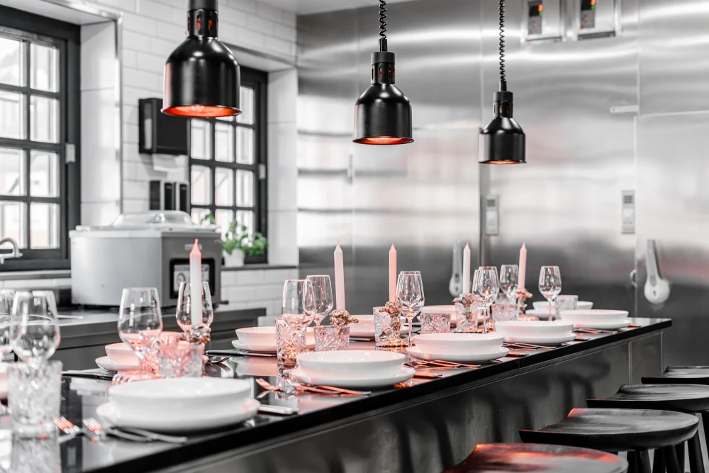 Gedeckter Tisch f&uuml;r ein Abendessen mit wei&szlig;en Tellern und Weingl&auml;sern unter schwarzen Pendelleuchten in einer modernen Edelstahl-K&uuml;che mit Theke und Industriedesign-Atmosph&auml;re.