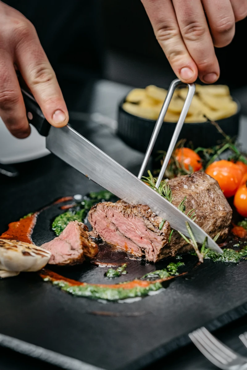 Zartes, rosa gebratenes Rindfleisch wird mit einer Gabel und einem Messer auf einem dunklen Teller angerichtet, umgeben von gebratenen Tomaten, Kr&auml;utern und einem Saucenspiegel.