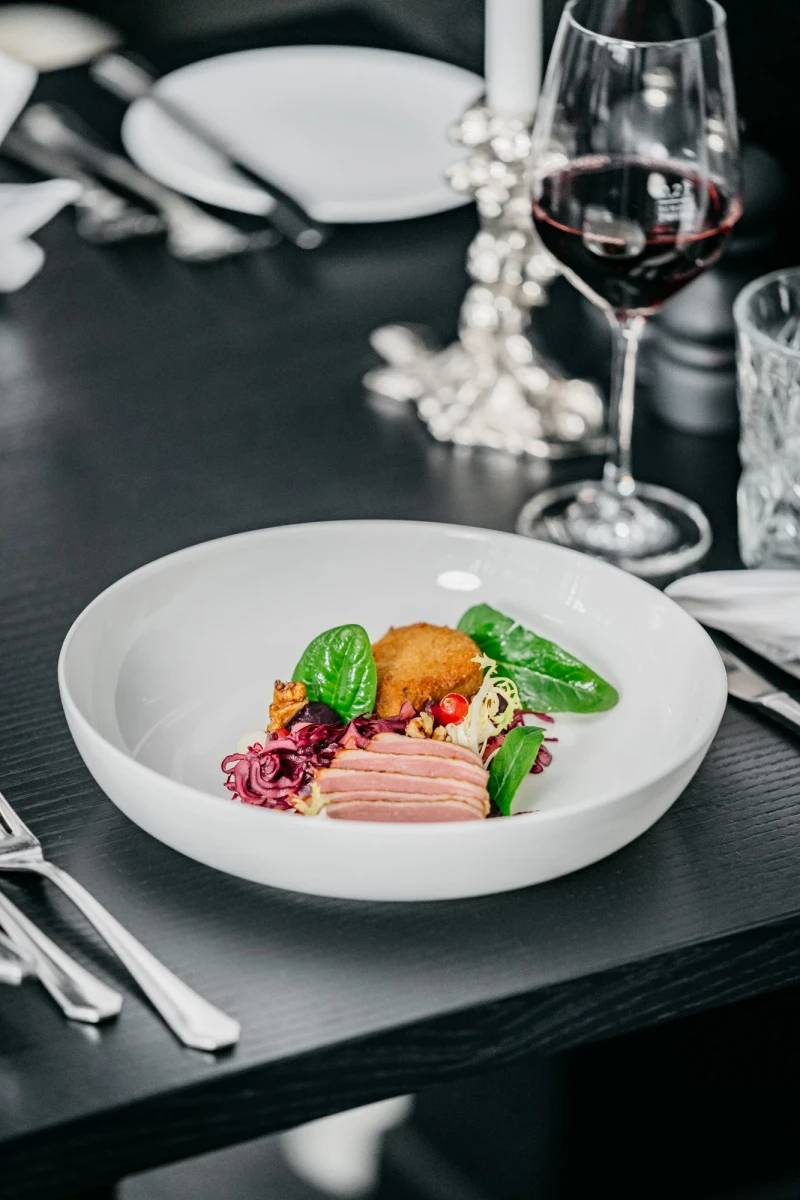 Elegante Fleischplatte mit zartem Schinken, einer orangefarbenen Zitrusfrucht und gr&uuml;nem Basilikum auf einem wei&szlig;en Teller, daneben ein Glas Rotwein und Silberbesteck auf dunklem Untergrund.