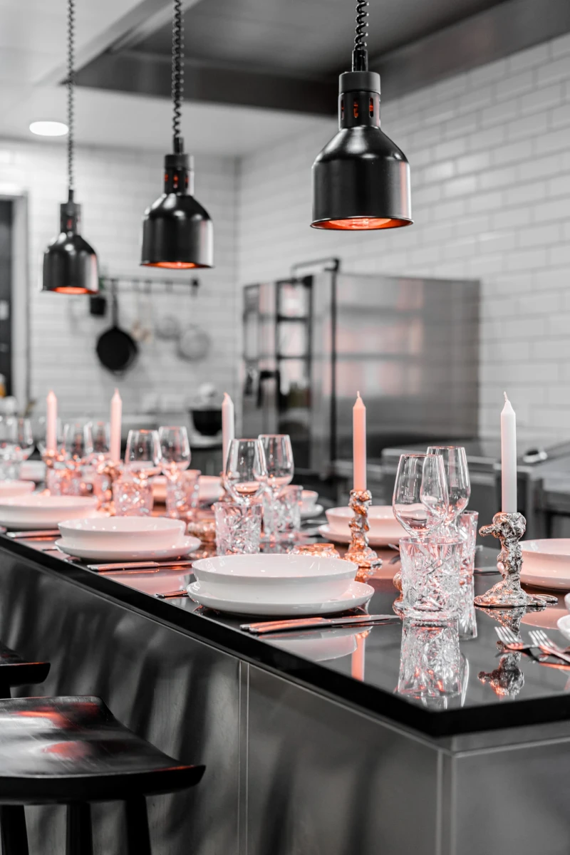 Elegant gedeckter Tisch in moderner Restaurantk&uuml;che mit schwarzen H&auml;ngelampen, wei&szlig;em Geschirr und Kerzen. Kristallgl&auml;ser und Dekoration in Pastellrosa auf einer dunklen Theke, die eine stilvolle Tischdekoration betont.
