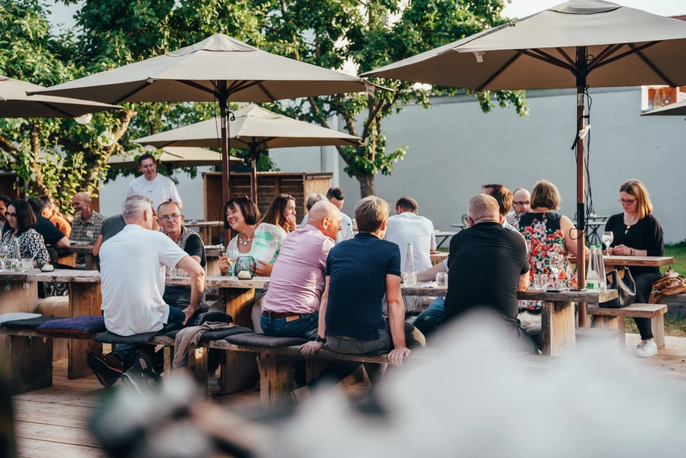 Mehrere Personen sitzen an Holzb&auml;nken unter wei&szlig;en Sonnenschirmen in einem Garten oder auf einer Terrasse. Die Atmosph&auml;re ist entspannt und sommerlich, mit B&auml;umen im Hintergrund. Verschiedene Altersgruppen unterhalten sich bei einem Sommerfest.