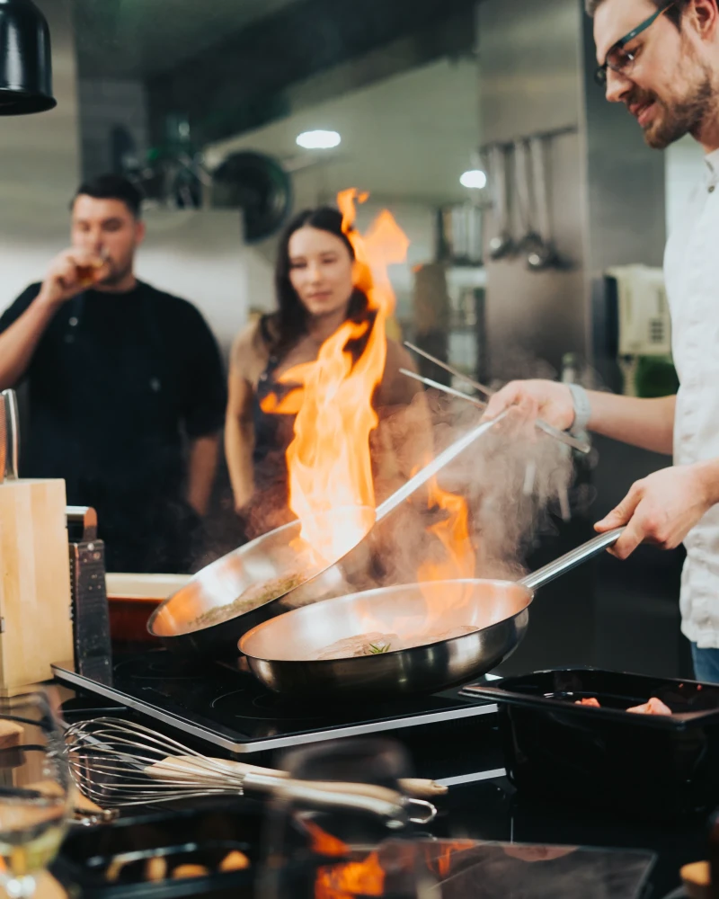 Profikoch flambiert mit hoher Flamme in einer professionellen Restaurantk&uuml;che. Zwei Kollegen beobachten die spektakul&auml;re Kochszene mit konzentriertem Blick. Kochutensilien und moderne K&uuml;chenausstattung sind im Vordergrund sichtbar.