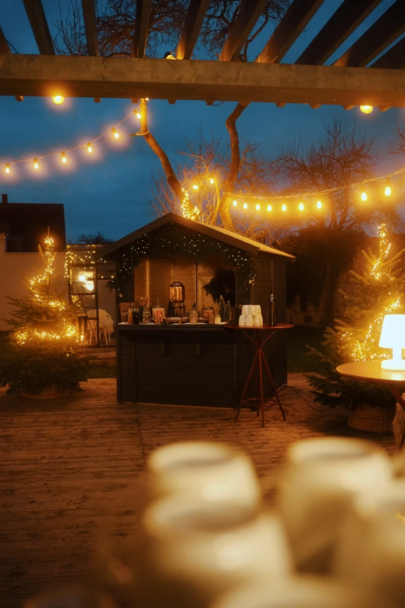 Beleuchteter Weihnachtsmarktstand mit warmen Lichterketten in einer gem&uuml;tlichen Winterabendatmosph&auml;re. Holzpergola und Verkaufsstand sind in goldene Lichter getaucht, umgeben von Weihnachtsb&auml;umen und einer stimmungsvollen Abendkulisse.