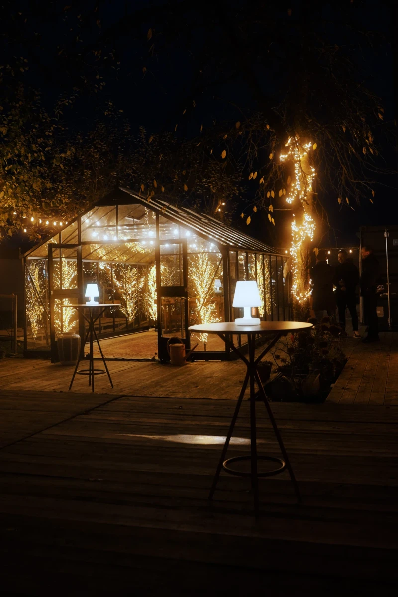 Beleuchtetes Gew&auml;chshaus in der Nacht mit warmen Lichterketten, umgeben von dunklen B&auml;umen. Zwei Stehlampen und Tische im Vordergrund auf einer Holzterrasse, die eine gem&uuml;tliche und romantische Atmosph&auml;re ausstrahlen.