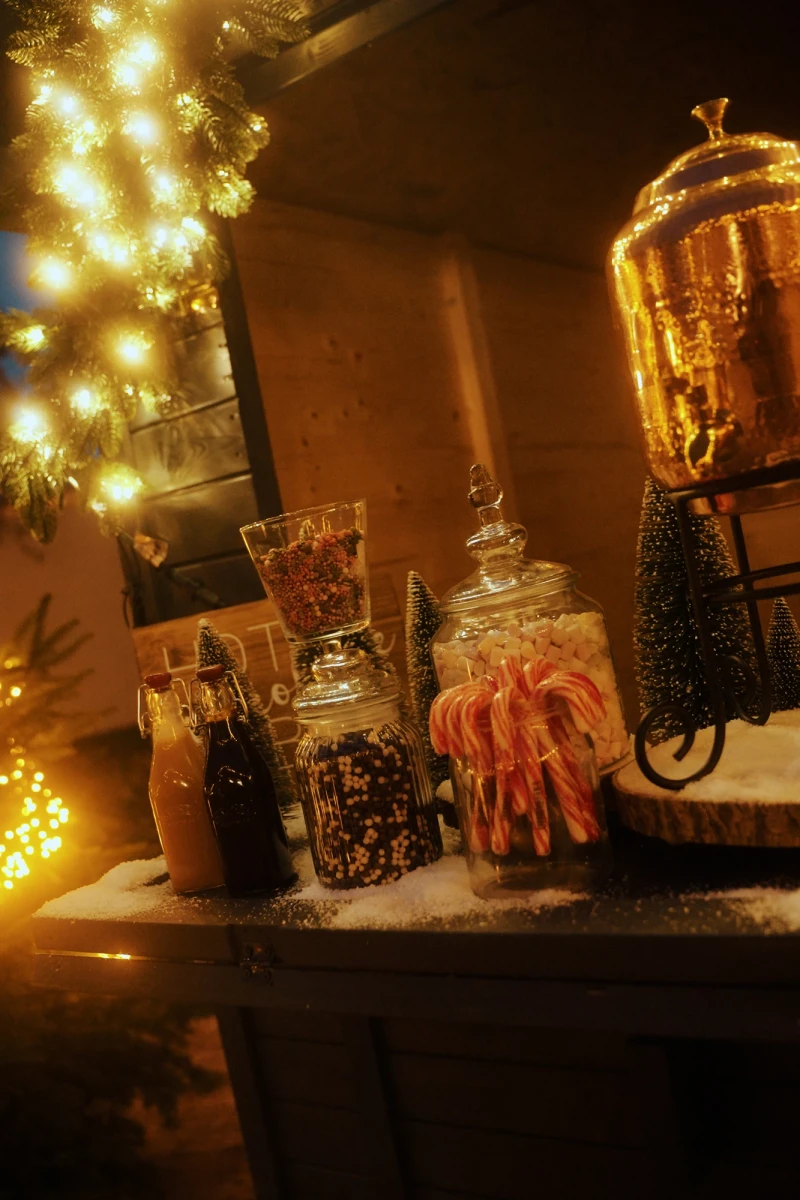 Weihnachtlich beleuchtete Theke mit Glasgef&auml;&szlig;en voller Kaffee, S&uuml;&szlig;igkeiten und Zimtstangen, im Hintergrund ein beleuchteter Weihnachtsbaum in warmen, goldenen T&ouml;nen.