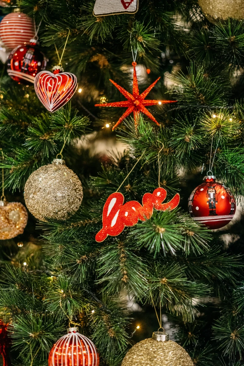Nahaufnahme eines Weihnachtsbaums mit verschiedenen Christbaumkugeln in Gold und Rot, darunter eine herzf&ouml;rmige Kugel, eine Schneeflocke und eine rote Schriftzug "Love". Die Kugeln h&auml;ngen an den gr&uuml;nen Zweigen des Tannenbaums.