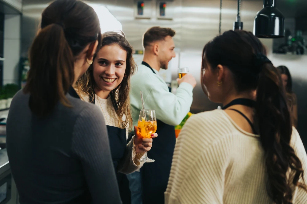 Drei junge Frauen unterhalten sich bei einer Feier in einem Raum, eine l&auml;chelt und h&auml;lt ein orangefarbenes Getr&auml;nk. Im Hintergrund steht eine weitere Person, die sich abwendet.