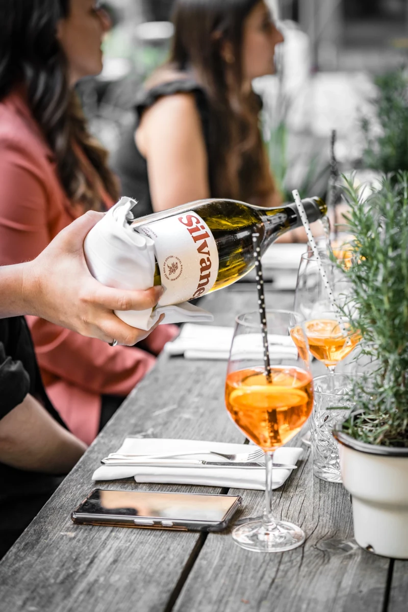Hand gie&szlig;t einen orangefarbenen Cocktail aus einer Flasche in ein Weinglas auf einem grauen Holztisch, im Hintergrund sitzen Menschen an einem Tisch mit Rosmarin-Pflanze.