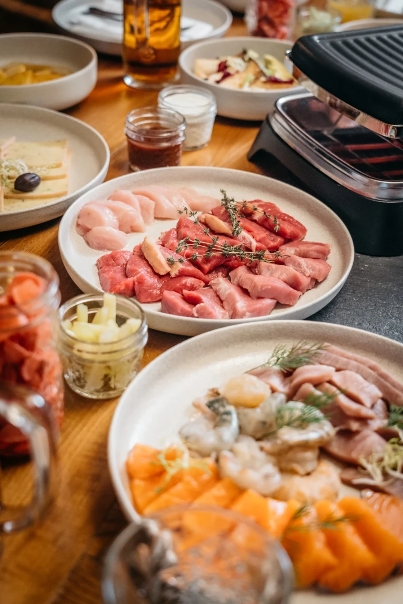 Verschiedene Teller mit Aufschnitt, Fleisch und Wurstwaren in Pastellfarben, dekoriert mit Kr&auml;utern. Ein Raclette- oder Fondue-Setup mit verschiedenen Fleischsorten und Beilagen auf einem Holztisch.