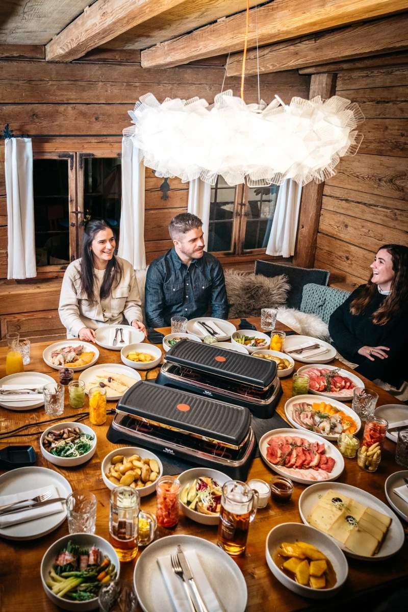 Drei Personen sitzen an einem Holztisch mit reichlich gedeckter Raclette-Grillplatte und verschiedenen Zutaten. Eine weiche Wolkenlampe h&auml;ngt &uuml;ber dem gem&uuml;tlichen Holztisch in einer rustikalen Holzh&uuml;tte, w&auml;hrend die Gruppe bereit ist, gemeinsam zu essen.