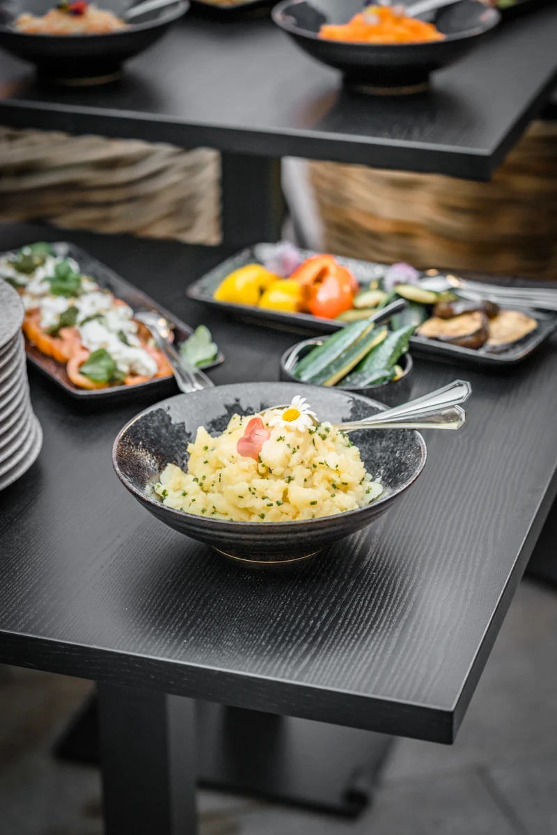 Gelber Kartoffelsalat in einer grauen Schale auf einem schwarzen Holztisch, umgeben von weiteren Tellerchen mit buntem Gem&uuml;se. Ein Silberl&ouml;ffel liegt in der Sch&uuml;ssel.